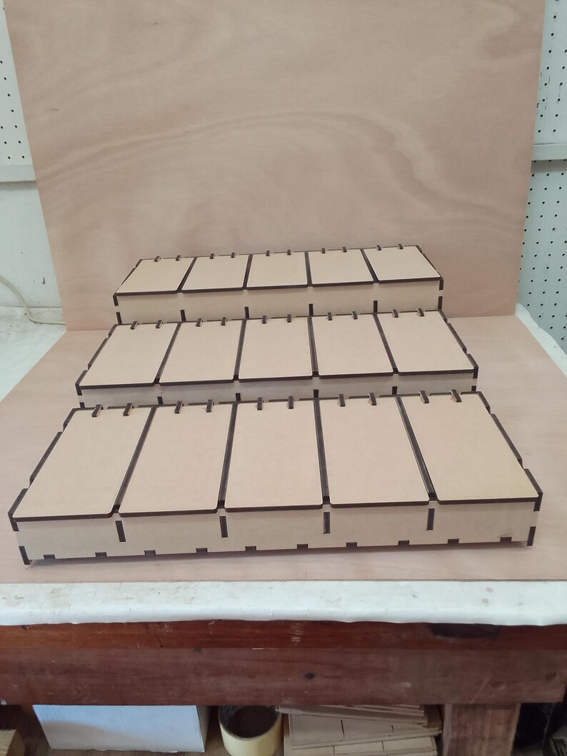 May include: Three-tiered, light brown wooden display shelves. Each shelf has multiple rectangular compartments. The shelves are arranged in a stepped configuration. The top shelf is the smallest, the bottom the largest.