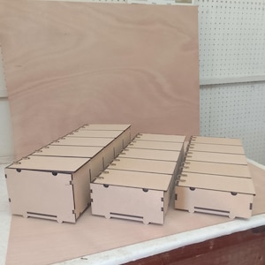 以下が含まれることがあります： さまざまなサイズの未完成の木製収納ボックスが3つ。各ボックスには、等間隔の溝と小さなハンドルカットアウトが付いた蓋が付いています。ボックスは明るい色の木材でできており、明るい茶色の木製の表面に配置されています。