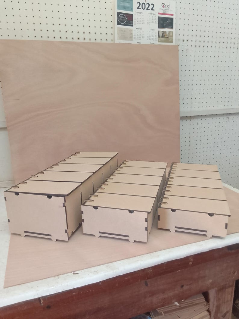 May include: Three unfinished wooden storage boxes of varying sizes. Each box has a lid with evenly spaced grooves and a small handle cutout. The boxes are made of light-coloured wood and are arranged on a light brown wooden surface.