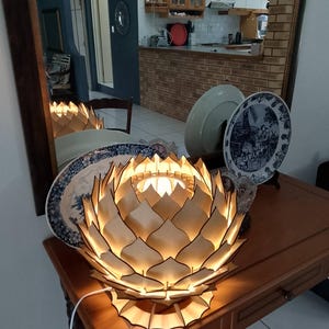 May include: A wooden lotus flower-shaped lamp with a warm glow sits on a wooden table. The lamp's petals are intricately cut, creating a layered design. A white power cord extends from the lamp. Decorative plates are in the background.