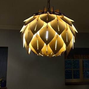 May include: A decorative pendant light fixture with a layered, leaf-like design. The light is made of a light-colored material, possibly wood, and is suspended from a chain. The fixture emits a warm, inviting glow, creating a focal point in the room.