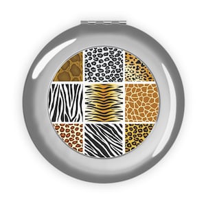 Può includere: Un specchietto compatto argentato con un motivo quadrato di stampe animalier, tra cui motivi zebrati, leopardati e giraffa. Lo specchietto è rotondo e ha una finitura metallica lucida.