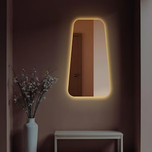 May include: A modern, rectangular mirror with a warm, white light around the perimeter. The mirror is mounted on a light brown wall, and there is a white table in the foreground.