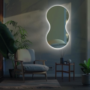 May include: A modern, oval-shaped mirror with a white LED light around the perimeter. The mirror is mounted on a wall in a room with a chair, a side table, and plants.