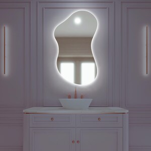 Peut inclure: Une salle de bain moderne avec un miroir de forme organique éclairé par des LED intégrées. La vanité comprend un lavabo blanc, une robinetterie cuivrée et un meuble blanc avec des détails dorés. Deux appliques murales verticales à LED encadrent le miroir.