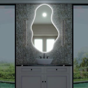 May include: A modern bathroom vanity with a white countertop and a white cabinet with two drawers. A white porcelain sink sits on the countertop, and a black faucet is mounted above it. A large, irregularly shaped mirror with a white LED light around the perimeter hangs on the wall behind the sink. The mirror reflects a view of a window with a green lawn and a pool outside.
