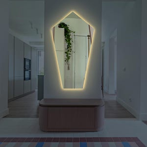 May include: A modern, geometric-shaped mirror with a warm white LED light border. The mirror is mounted on a white wall above a wooden bench.