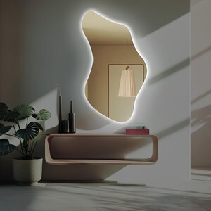 May include: A modern, irregularly shaped mirror with a white LED light around the perimeter. The mirror is mounted on a white wall above a light wood console table with a shelf. There are two black candlesticks on the table.