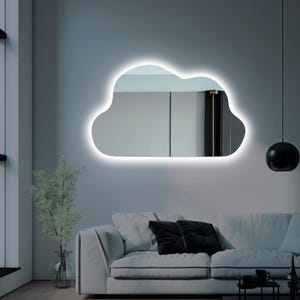 以下が含まれることがあります： 明るい白いLEDの輪郭を持つ雲型の鏡で、灰色の壁に取り付けられています。鏡は白いソファ、黒いクッション、黒いペンダントライトのある部屋を映しています。左側には緑の入った花瓶があります。