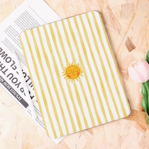 May include: A rectangular case with a yellow and white striped pattern and a smiling sun illustration. The case is on a light-colored wooden surface, with a newspaper and a pink tulip in the background.