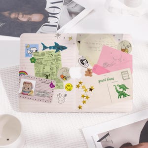 Op de afbeelding: Een roze laptop met verschillende stickers, waaronder een blauwe walvis, een gele smiley, een groene dinosaurus en een roze slak. De laptop staat op een wit oppervlak met een tijdschrift en een witte pot.