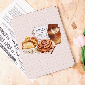 May include: A light pink tablet case featuring illustrations of cafe treats. The design includes a cinnamon roll, a sandwich, a coffee drink, and a receipt from "Donut Cafe." The background has a grid pattern.