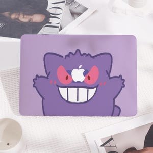 May include: A purple laptop case featuring a cartoon ghost-like character with a wide grin and red eyes. The character has an Apple logo in place of its eyes. The case is designed to fit a laptop.