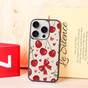 Puede incluir: Una funda de teléfono con un diseño de cerezas. La funda presenta un fondo beige claro con cerezas rojas, hojas verdes y pequeños corazones rojos. También hay un lazo rojo. Las lentes de la cámara del teléfono son visibles.