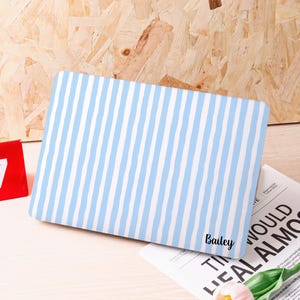 Może przedstawiać: Prostokątne etui na laptopa w niebiesko-białe paski. Imię "Bailey" jest wydrukowane czcionką na etui. Etui leży na jasnej powierzchni. Tło ma drewnianą fakturę.