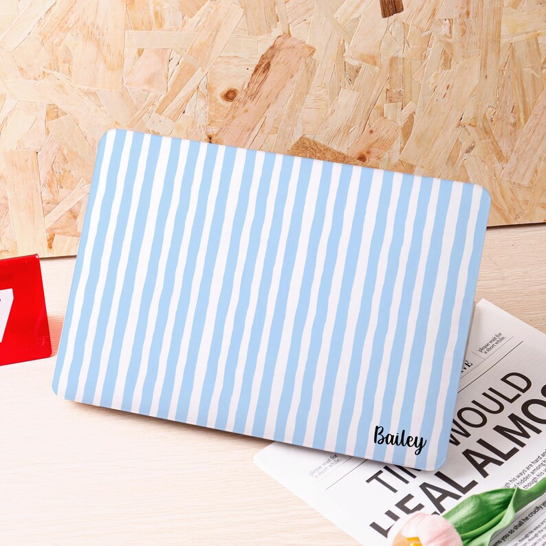 Może przedstawiać: Prostokątne etui na laptopa w niebiesko-białe paski. Imię "Bailey" jest wydrukowane czcionką na etui. Etui leży na jasnej powierzchni. Tło ma drewnianą fakturę.
