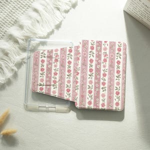 May include: A clear phone case with a floral patterned phone wallet. The wallet features vertical stripes of pink and white with pink flowers and green leaves. The case and wallet are on a white surface.