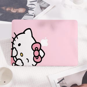 May include: A pink laptop case featuring a large graphic of Hello Kitty. The white Hello Kitty character has black eyes, whiskers, and a pink bow. The Apple logo is visible on the case.