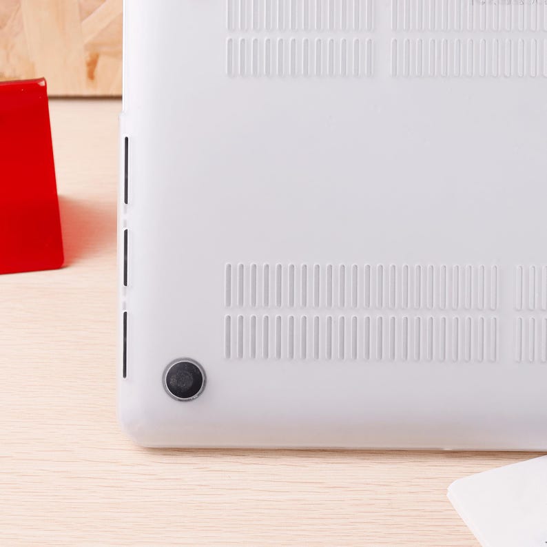 Może przedstawiać: Białe etui na laptopa z otworami wentylacyjnymi i czarną gumową n&oacute;żką. Obudowa ma gładką powierzchnię i ma chronić laptopa. Obudowa znajduje się na jasnej drewnianej powierzchni.