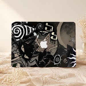 May include: A laptop skin featuring a black and white collage design with a central star shape surrounding an Apple logo. The graphic includes a stylized cat, swirling patterns, musical notes, a crescent moon, an eye, and various star motifs.
