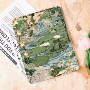 Op de afbeelding: Een decoratieve tablethoes met een serene vijverscène, met een kikker op een waterlelie, witte bloemen en een vogel. Het ontwerp bevat het woord "healing" en tekst uit een krant. Vintage, botanische esthetiek.