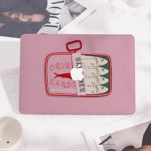 Puede incluir: Una funda rosa para portátil con un diseño de lata de sardinas. La ilustración de la lata incluye las palabras "ORIGINAL SARDINES WITH TOMATO SAUCE" y muestra cuatro peces. La funda tiene una lengüeta roja y un logotipo blanco de Apple.