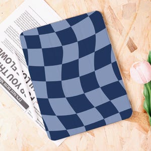 Può includere: Una custodia per tablet con un motivo a scacchi blu navy e azzurro. Il motivo ha un design curvo e ondulato. La custodia è su una superficie di legno con un giornale e un fiore rosa.