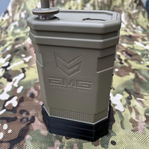 May include: An olive green, rectangular container with a black base and a top with a small, cylindrical knob. The container has the text "EMG INTERNATIONAL" and a logo on the front. The container is sitting on a camouflage patterned surface.