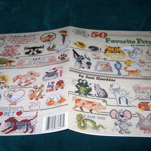 May include: A book titled "50 Favourite Pets to Cross Stitch" by Sam Hawkins. The book features a variety of colourful cross stitch patterns of animals, including cats, dogs, birds, and fish. The cover of the book is white with a red and blue border.