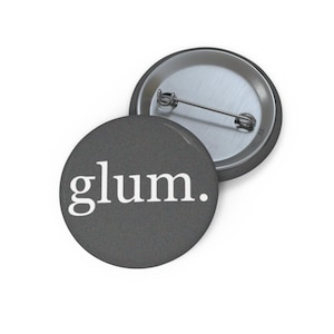 May include: Two round, gray pinback buttons. One button displays the word "glum." in white, sans-serif font. The other button is silver and shows the pin mechanism. The buttons are on a white background.