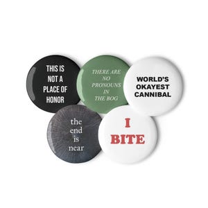 Può includere: Cinque spille rotonde con testi diversi. Una spilla nera riporta THIS IS NOT A PLACE OF HONOR, una verde dice THERE ARE NO PRONOUNS IN THE BOG, una bianca dice WORLD'S OKAYEST CANNIBAL, una scura testurizzata dice the end is near e una bianca dice I BITE.