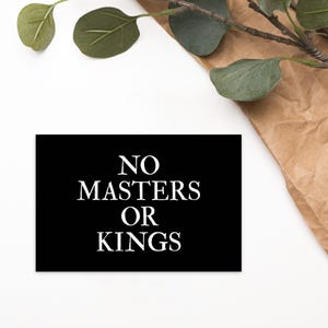 May include: A rectangular black sign featuring the white text NO MASTERS OR KINGS centered in a clean, bold serif font. The sign is presented against a plain white background with a decorative sprig of green eucalyptus leaves and brown paper.