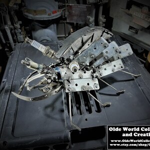 May include: A metal sculpture of a beetle made from recycled materials, including spark plugs. The sculpture is on a dark gray surface. The sculpture is for sale by Olde World Collectibles and Creations.