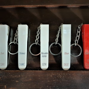 May include: A set of six white and red keychains with different musical instrument names printed on them. The keychains are labeled Sax, Chimes, Hawaiian Guitar, Oboe, Clarinet, and 16 Clar.
