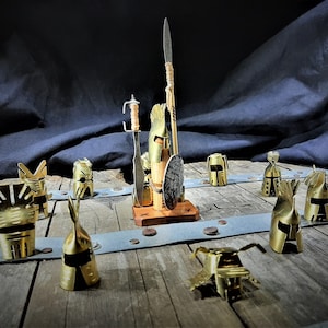 Op de afbeelding: Een verzameling miniatuur metalen helmen, sommige met zwaarden en schilden, gerangschikt op een houten oppervlak. De helmen zijn goudkleurig en hebben ingewikkelde ontwerpen.