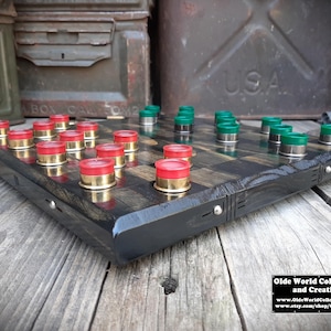 May include: A wooden checkerboard game with red and green shotgun shell caps as playing pieces. The board is made of dark wood with a black stain and has a distressed look. The board is sitting on a wooden surface.