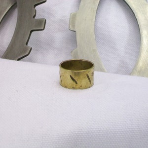 Può includere: Un anello di metallo dorato con una superficie testurizzata. L'anello è appoggiato su una superficie di tessuto bianco.