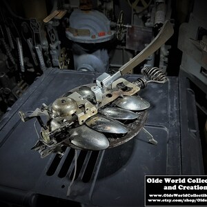 May include: A whimsical metal sculpture made from recycled spoons, bells, and other metal objects. The sculpture is shaped like a bug with a long, thin body and a pair of wings. It has a lever on the back that can be used to make the wings move.