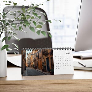 Könnte beinhalten: Ein Tischkalender für Juni mit einem Foto einer europäischen Straßenszene. Der Kalender ist spiralgebunden und steht auf einem Holzschreibtisch. Ein Computermonitor und Grünzeug sind im Hintergrund zu sehen.