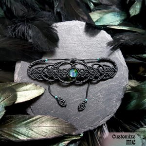 Könnte beinhalten: Schwarzes Makramee-Armband mit einem grün schimmernden Edelstein in der Mitte. Das Armband hat ein geknüpftes Design und ist mit türkisfarbenen Perlen verziert.