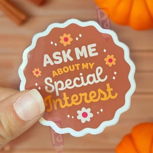 May include: A round sticker with a scalloped white border and the text "ASK ME ABOUT MY Special Interest" in a mix of brown and yellow. The sticker is decorated with small flower and dot graphics. The background includes pumpkins and a wooden surface.