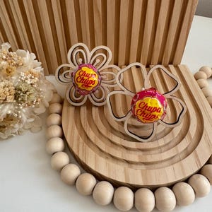 May include: Two wooden flower-shaped lollipop holders, each holding a Chupa Chups lollipop. The holders are on a wooden tray with a beaded garland and a wooden background. The lollipops have pink and yellow wrappers.