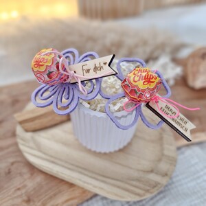 May include: A white cupcake-shaped container with decorative purple flower cutouts holding two Chupa Chups lollipops. Each lollipop is attached to a small wooden tag with German text. The container sits on a wooden tray.