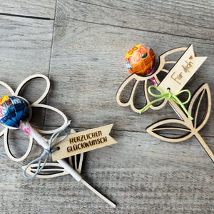 May include: Two wooden flower-shaped lollipop holders. One has a blue and yellow lollipop with a banner that reads "HERZLICHEN GL&Uuml;CKWUNSCH". The other has an orange lollipop and a banner that reads "For dich".