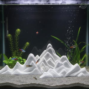 May include: A white, mountain-shaped aquarium decoration. The decor is made of a textured material and is placed in an aquarium with fish and aquatic plants. The background is dark, and bubbles rise from the bottom.