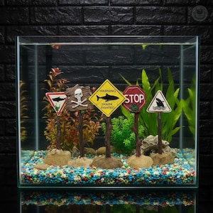 Puede incluir: Un conjunto de cinco letreros decorativos para acuario que incluyen una señal triangular de advertencia de tiburón, una calavera, un rombo amarillo con el texto WARNING SHARK SIGHTED, una señal octogonal roja de STOP y una señal triangular.