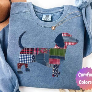 May include: Light blue Comfort Colors sweatshirt featuring a patchwork dachshund design. The dog's body is composed of various fabric patterns, including plaid and floral prints. The sweatshirt has a crew neck and the Comfort Colors logo.