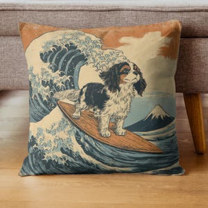 May include: Square throw pillow featuring a Cavalier King Charles Spaniel surfing on a wooden surfboard in front of a large ocean wave. The design is inspired by Japanese art, with a mountain in the background. The color palette includes shades of blue, orange, and brown.