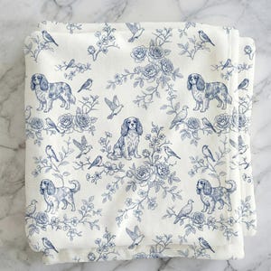 May include: Folded white fabric with a blue toile pattern featuring Cavalier King Charles Spaniels, birds, and floral elements. The design includes roses and branches, creating a classic, decorative aesthetic.