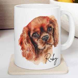 Peut inclure: Mug en céramique blanche avec un portrait à l'aquarelle d'un Cavalier King Charles, avec une fourrure marron et rousse. Le nom du chien, "Ruby", est écrit en cursive. La tasse a une anse blanche et repose sur un dessous de verre beige.